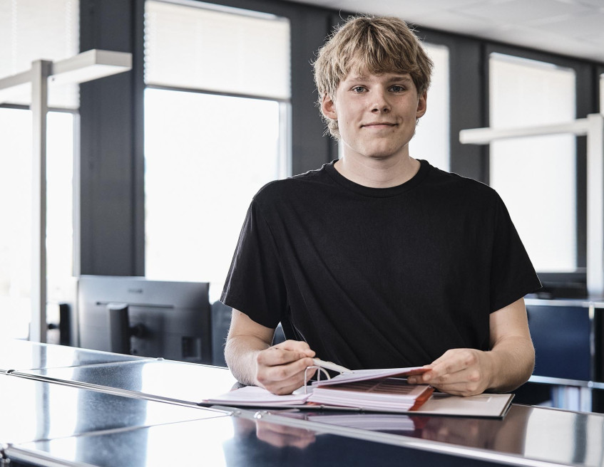 Lernender der emmental versicherung steht vor seinem Arbeitspatz und blickt freundlich in die Kamera.