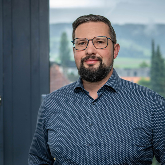 Kundenberater der emmental versicherung steht vor einem Fenster und blickt freundlich in die Kamera.