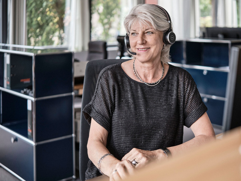 Eine Mitarbeiterin der emmental versicherung sitz an ihrem Arbeitsplatz und ist über ihr Headset am telefonieren.