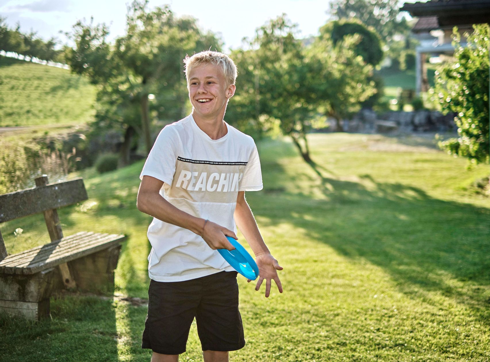 Ein blonder Junge steht an einem Sommertag im Garten und spielt Frisbee.