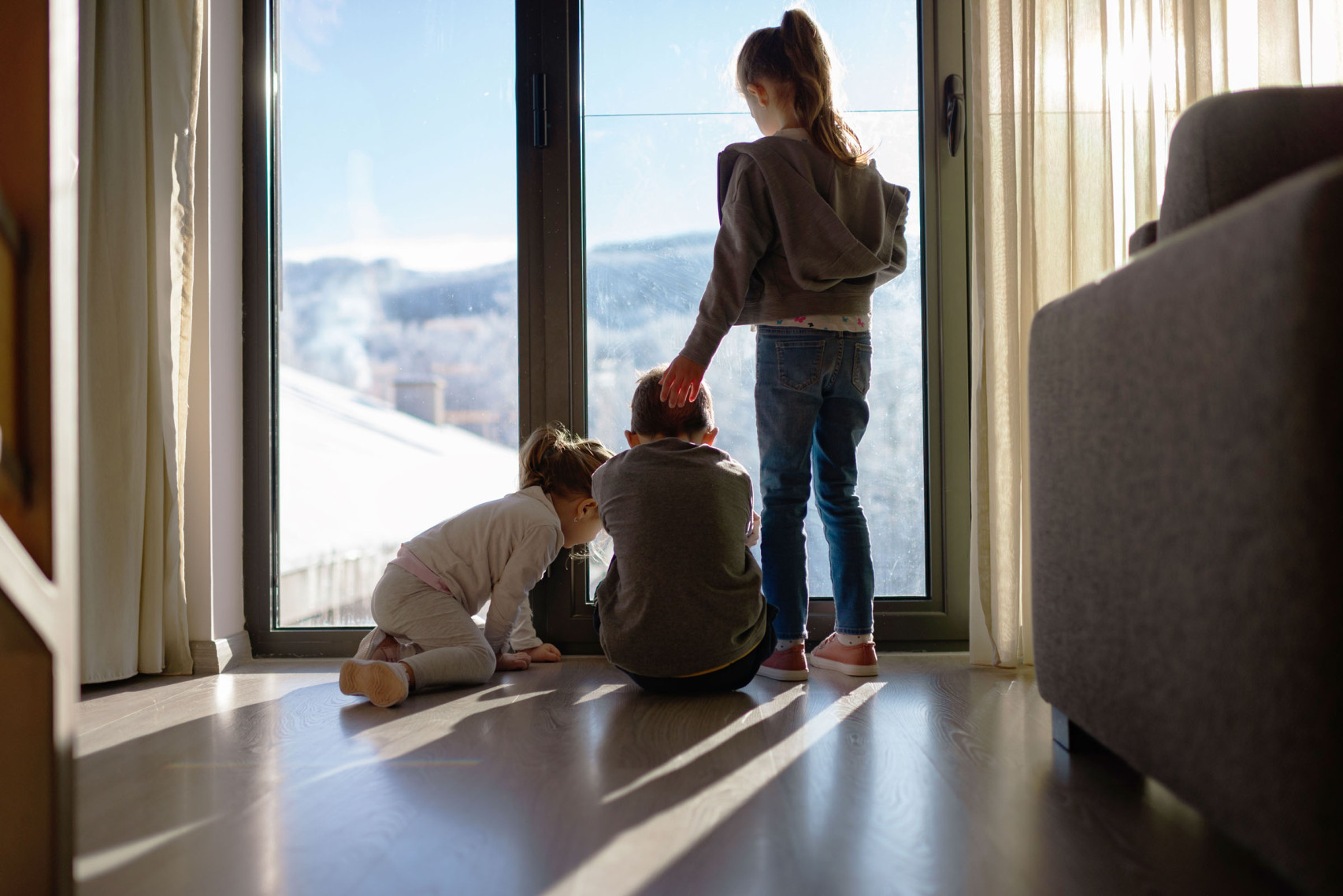 Drei junge Kindern stehen vor dem Fenster im Wohnzimmer und schauen hinaus.
